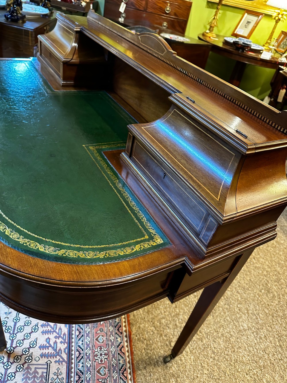 An antique mahogany writing desk featuring a green leather surface, a raised central superstructure with small drawers, satinwood string inlay, and tapered legs on brass castors. View VI