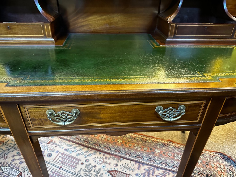An antique mahogany writing desk featuring a green leather surface, a raised central superstructure with small drawers, satinwood string inlay, and tapered legs on brass castors. View IX