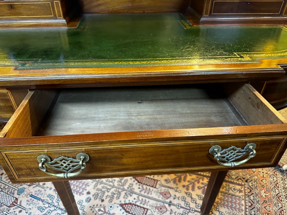 An antique mahogany writing desk featuring a green leather surface, a raised central superstructure with small drawers, satinwood string inlay, and tapered legs on brass castors. View X