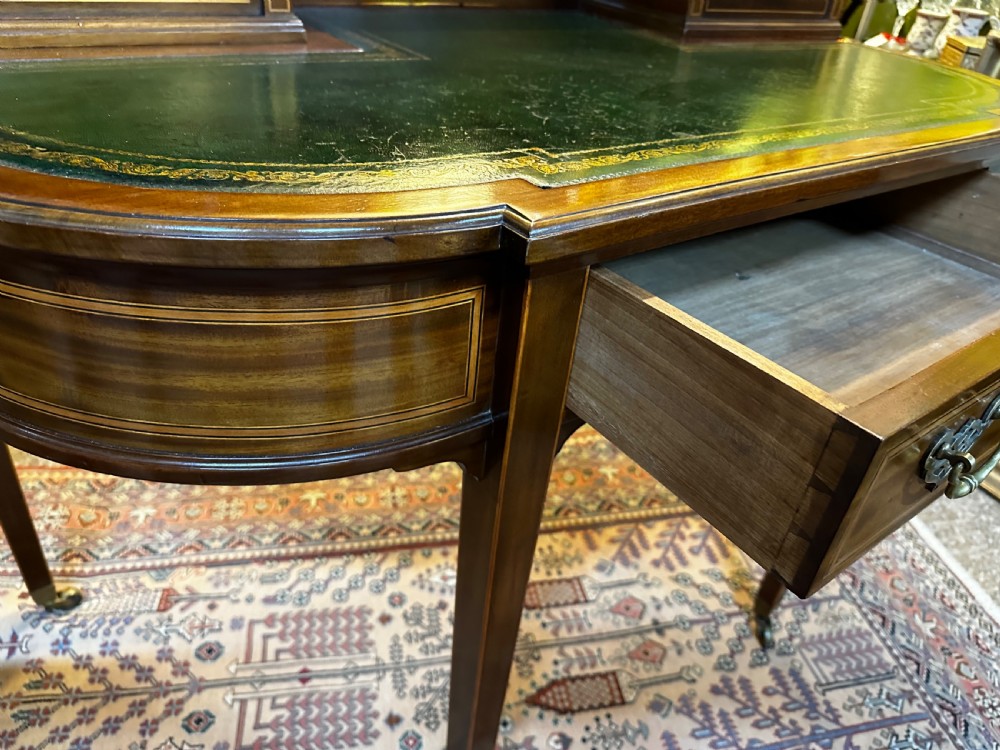 An antique mahogany writing desk featuring a green leather surface, a raised central superstructure with small drawers, satinwood string inlay, and tapered legs on brass castors. View XI