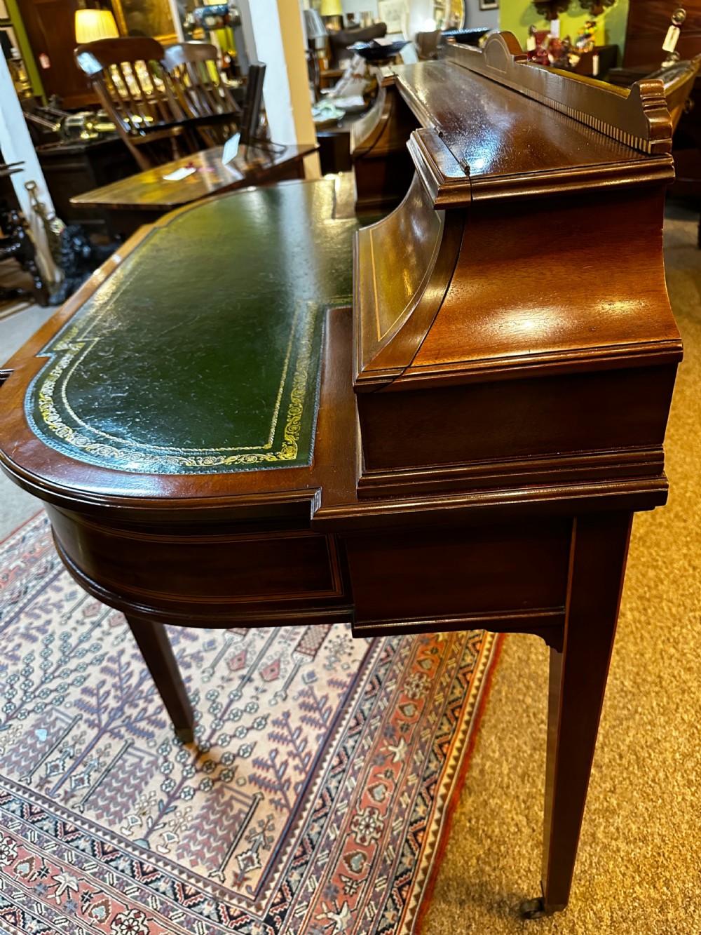 An antique mahogany writing desk featuring a green leather surface, a raised central superstructure with small drawers, satinwood string inlay, and tapered legs on brass castors. View XIII