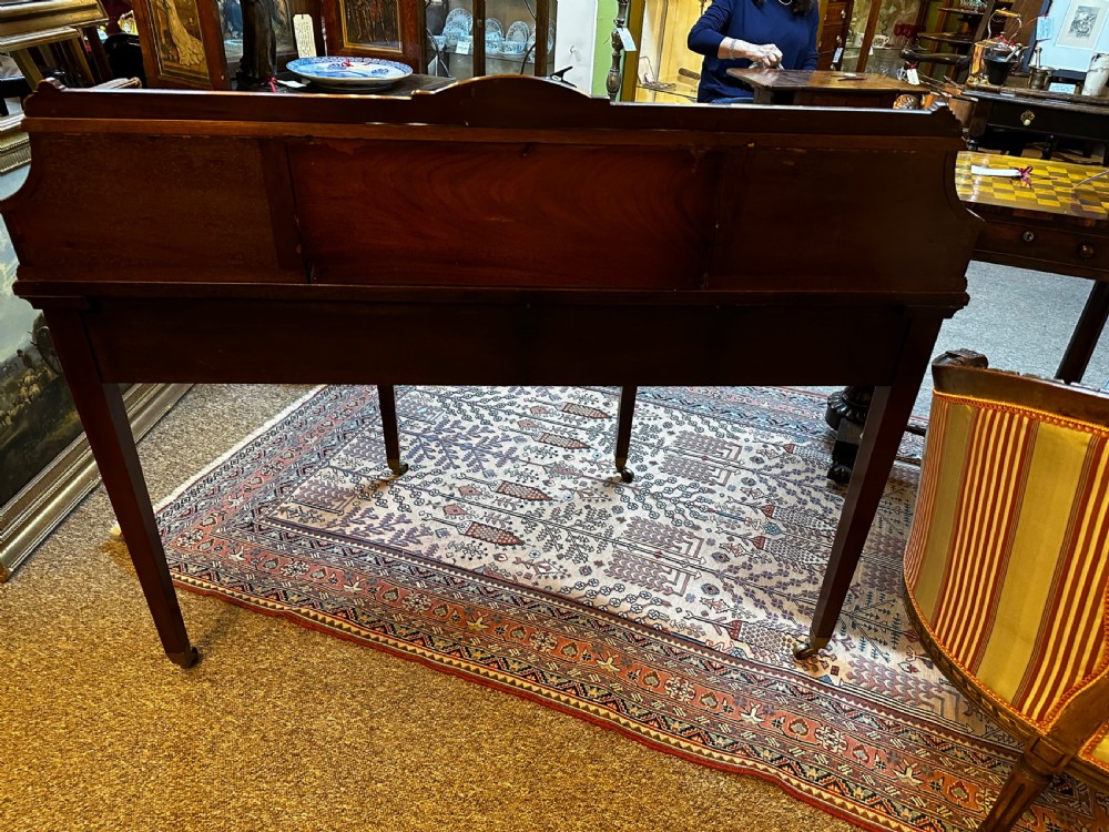 An antique mahogany writing desk featuring a green leather surface, a raised central superstructure with small drawers, satinwood string inlay, and tapered legs on brass castors. View XIV