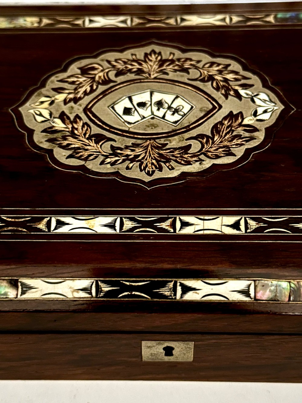 A rectangular dark rosewood games box featuring mother-of-pearl and abalone inlaid borders with a central playing card motif medallion and a brass keyhole. View XI