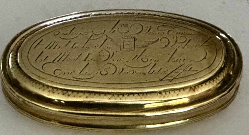 An 18th-century oval brass tobacco box featuring hand-engraved Dutch calligraphy and a hinged lid.
