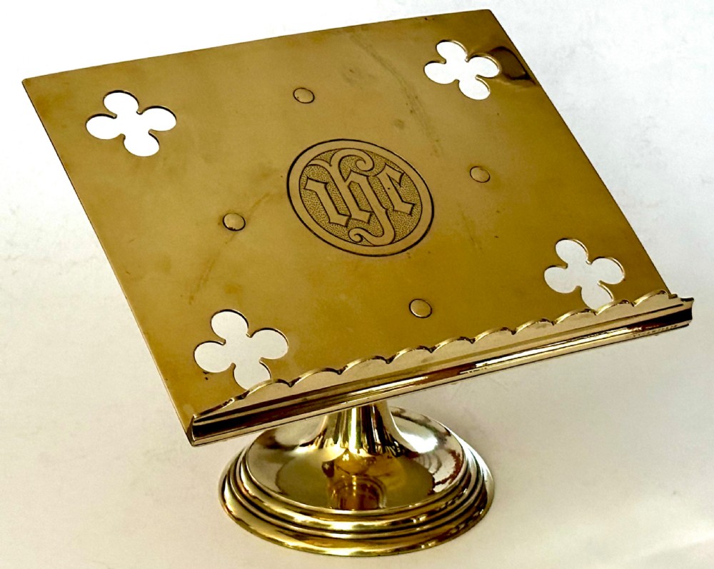 A polished brass tabletop lectern featuring quatrefoil corner cutouts, an engraved Gothic monogram inside a central medallion, a scalloped lip, and a stepped circular pedestal base.