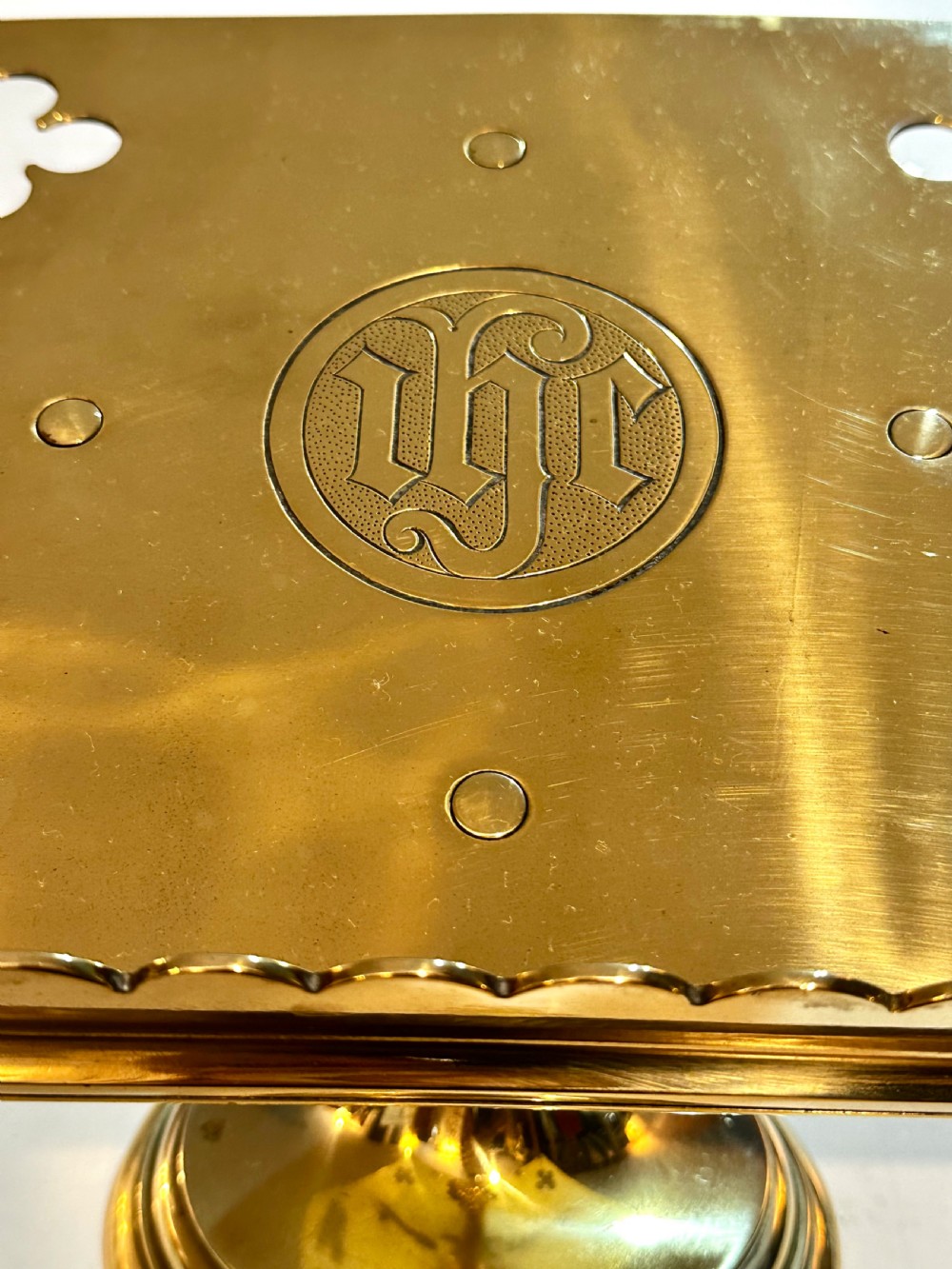 A polished brass tabletop lectern featuring quatrefoil corner cutouts, an engraved Gothic monogram inside a central medallion, a scalloped lip, and a stepped circular pedestal base. View VI