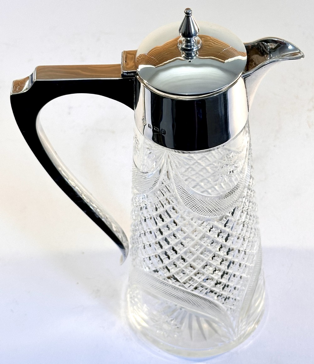 A tall cut glass claret jug decorated with diamond cut facets, featuring a polished silver-plated collar, hinged lid with finial, and an angular handle. View IV