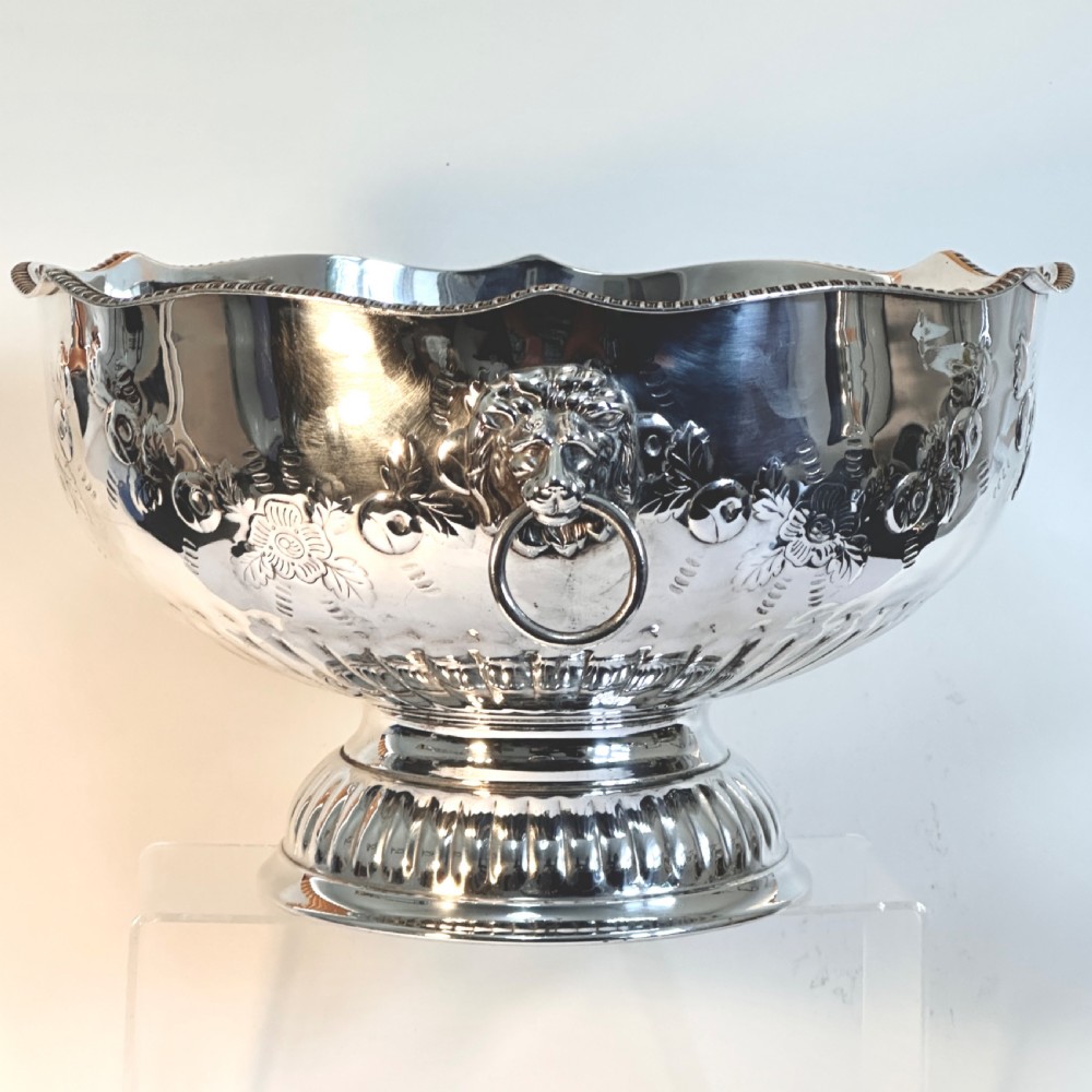 A silver-plated pedestal punch bowl featuring embossed floral repousse decoration, a scalloped rim, and twin lion mask drop-ring handles.
