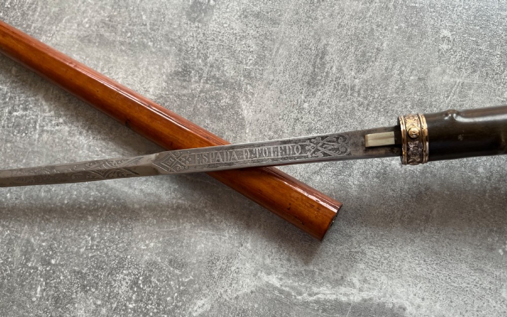 An antique malacca wood sword stick featuring a curved horn handle, decorative brass collar with push button release, and an etched steel Toledo blade. View IV