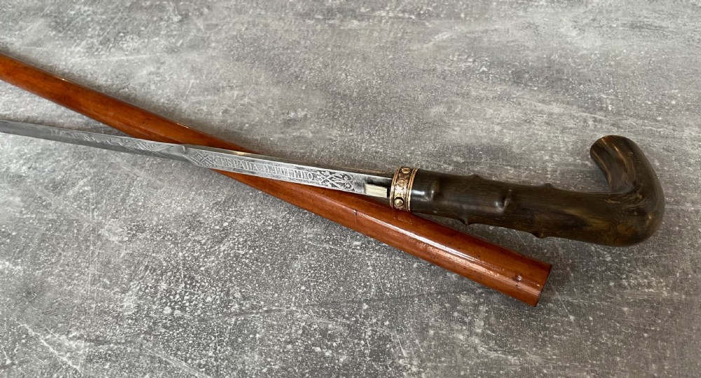 An antique malacca wood sword stick featuring a curved horn handle, decorative brass collar with push button release, and an etched steel Toledo blade. View V