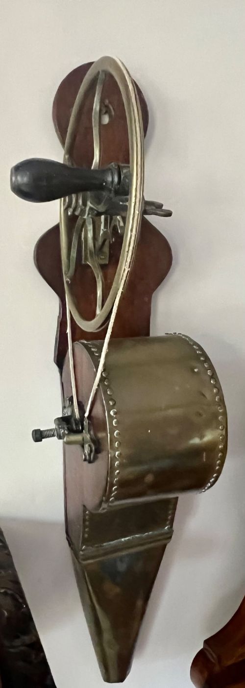 An antique mechanical rotary fireplace bellows featuring a mahogany wooden base, a brass spoked wheel with a hand crank, a driving band, and a brass casing leading to a tapered nozzle. View VI