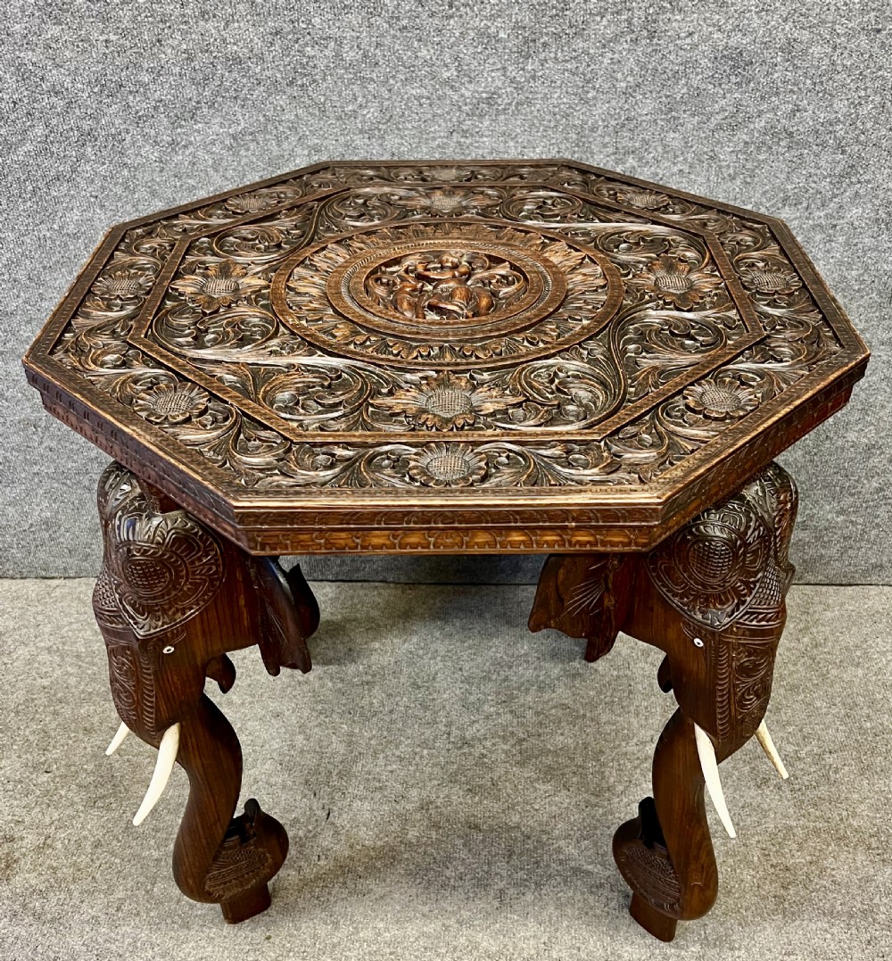 An octagonal dark brown teak coffee table supported by four intricately carved elephant legs with upturned trunks and tusks, featuring an elaborately carved floral top surface. View II
