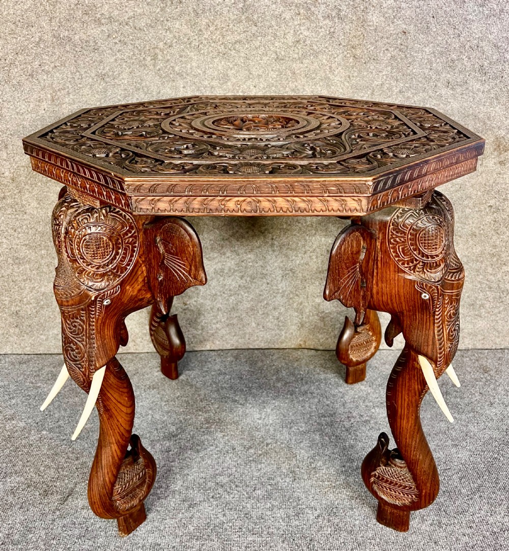 An octagonal dark brown teak coffee table supported by four intricately carved elephant legs with upturned trunks and tusks, featuring an elaborately carved floral top surface. View V