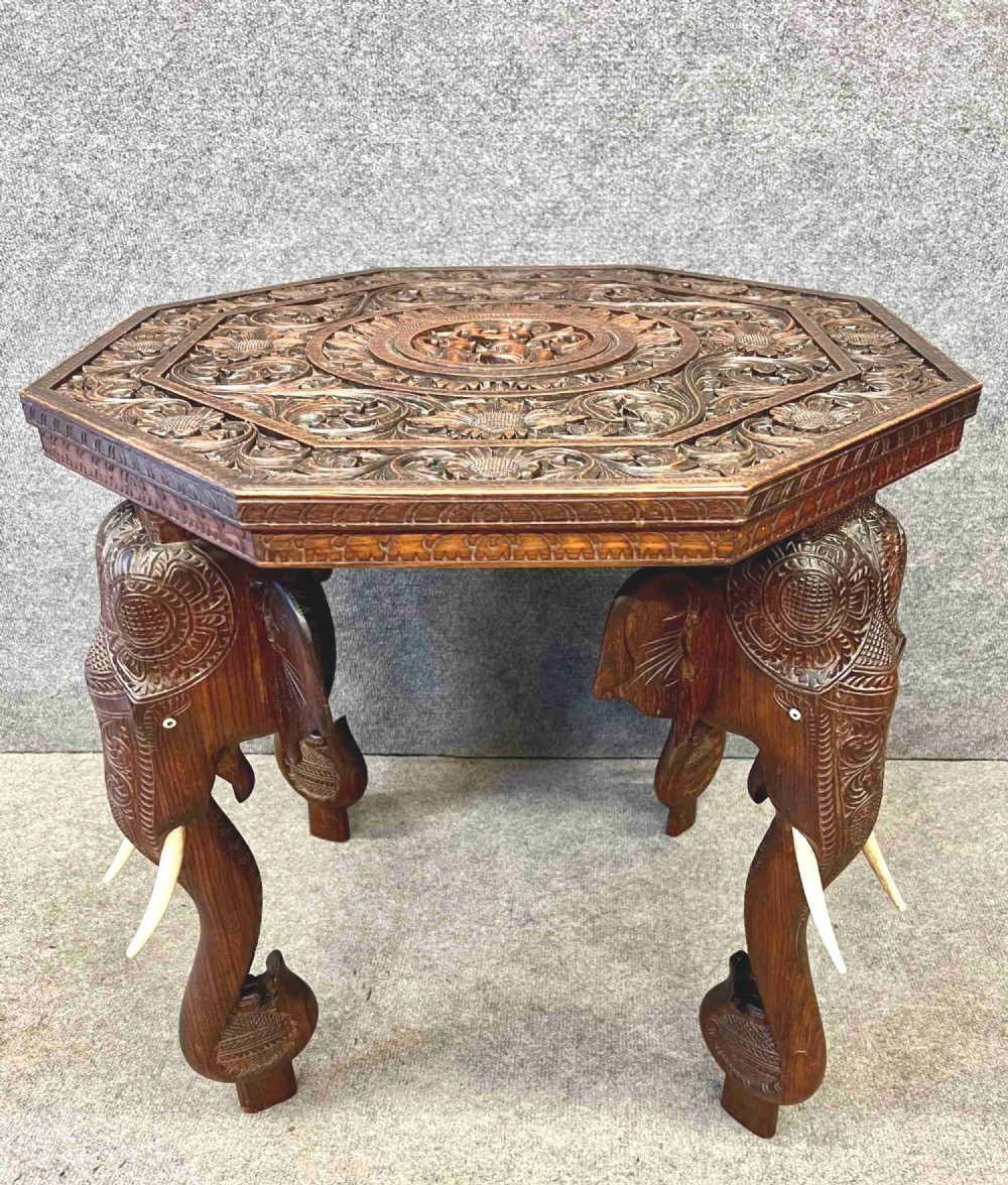 An octagonal dark brown teak coffee table supported by four intricately carved elephant legs with upturned trunks and tusks, featuring an elaborately carved floral top surface.