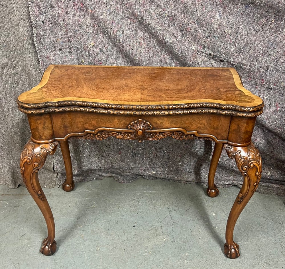 A burr walnut card table featuring a serpentine top with crossbanded borders, an ornate shell-carved apron, and cabriole legs ending in ball and claw feet.