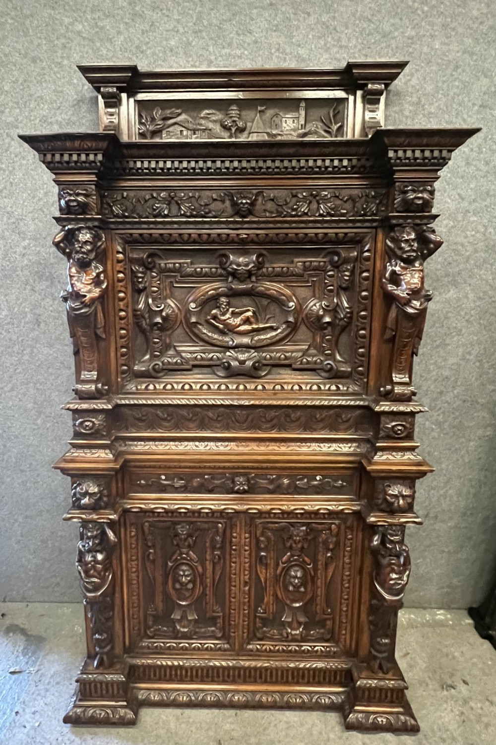 A heavily carved dark walnut cabinet featuring figurative reliefs, caryatid supports, mask motifs, and an architectural cresting.