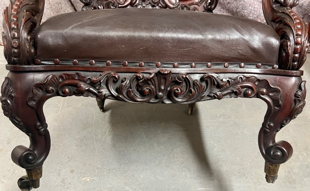 A Victorian carved mahogany armchair featuring deep-buttoned brown leather upholstery, elaborate scrollwork carving, carved armrests, and cabriole legs with brass castors. View II