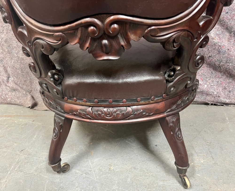 A Victorian carved mahogany armchair featuring deep-buttoned brown leather upholstery, elaborate scrollwork carving, carved armrests, and cabriole legs with brass castors. View IV