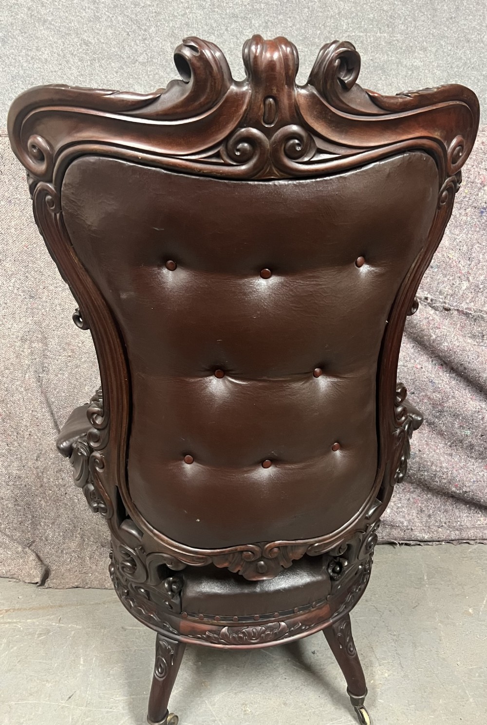 A Victorian carved mahogany armchair featuring deep-buttoned brown leather upholstery, elaborate scrollwork carving, carved armrests, and cabriole legs with brass castors. View V