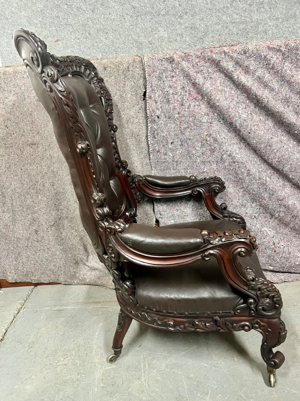 A Victorian carved mahogany armchair featuring deep-buttoned brown leather upholstery, elaborate scrollwork carving, carved armrests, and cabriole legs with brass castors. View VI