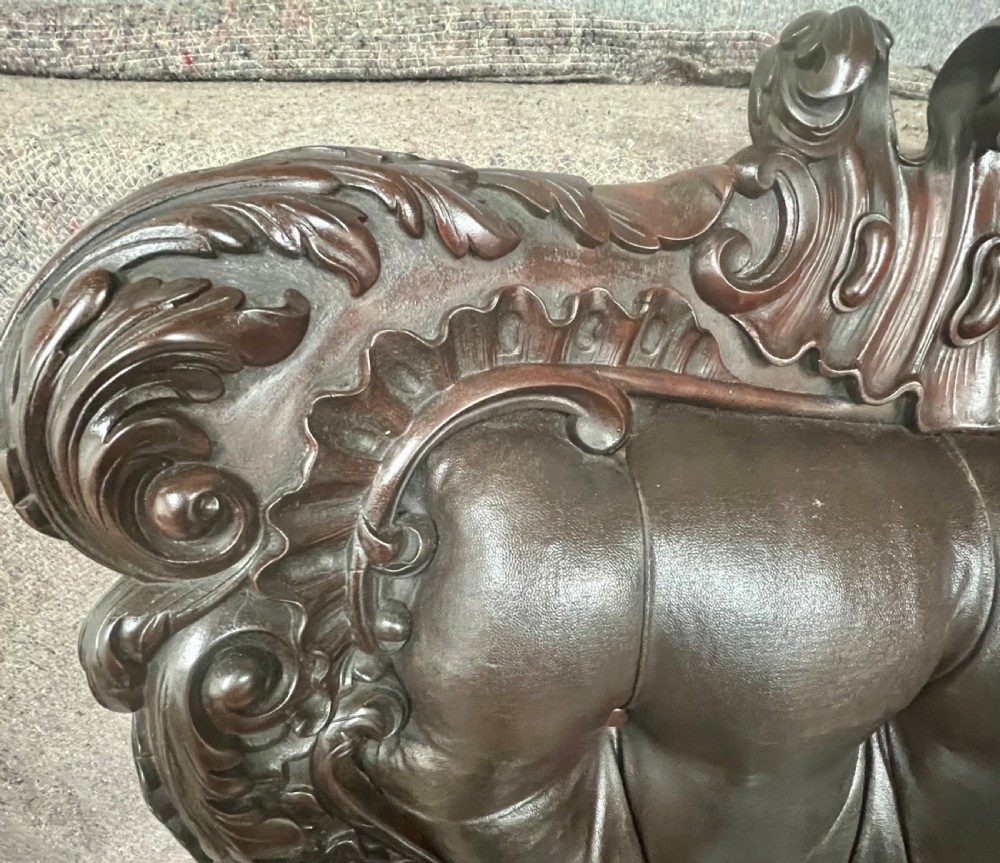 A Victorian carved mahogany armchair featuring deep-buttoned brown leather upholstery, elaborate scrollwork carving, carved armrests, and cabriole legs with brass castors. View VII