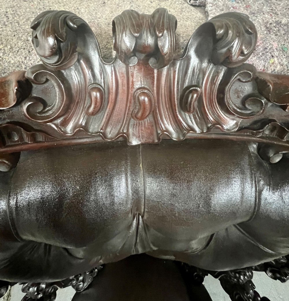 A Victorian carved mahogany armchair featuring deep-buttoned brown leather upholstery, elaborate scrollwork carving, carved armrests, and cabriole legs with brass castors. View VIII