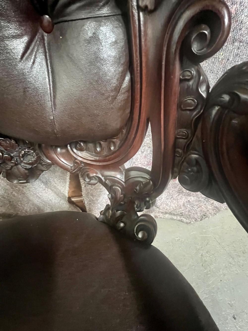 A Victorian carved mahogany armchair featuring deep-buttoned brown leather upholstery, elaborate scrollwork carving, carved armrests, and cabriole legs with brass castors. View X