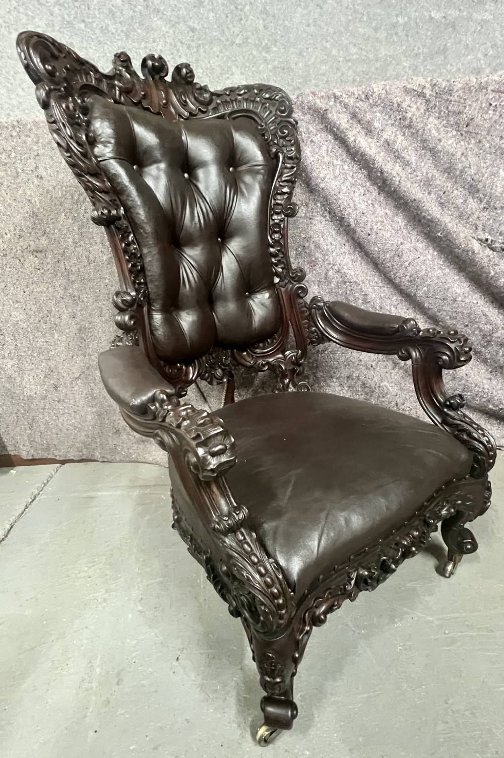 A Victorian carved mahogany armchair featuring deep-buttoned brown leather upholstery, elaborate scrollwork carving, carved armrests, and cabriole legs with brass castors. View XI