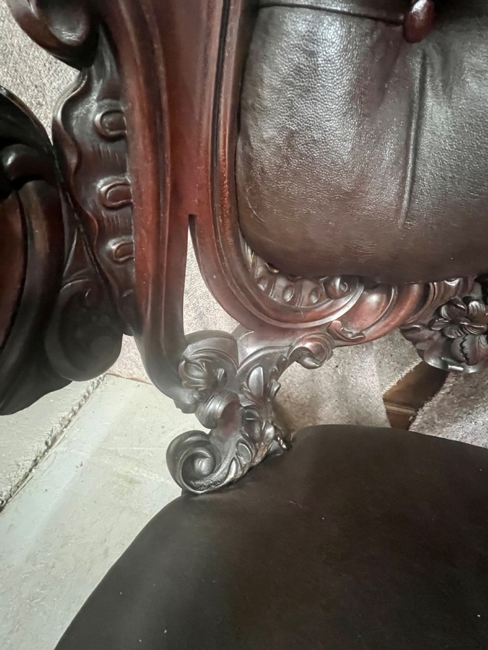 A Victorian carved mahogany armchair featuring deep-buttoned brown leather upholstery, elaborate scrollwork carving, carved armrests, and cabriole legs with brass castors. View XIII