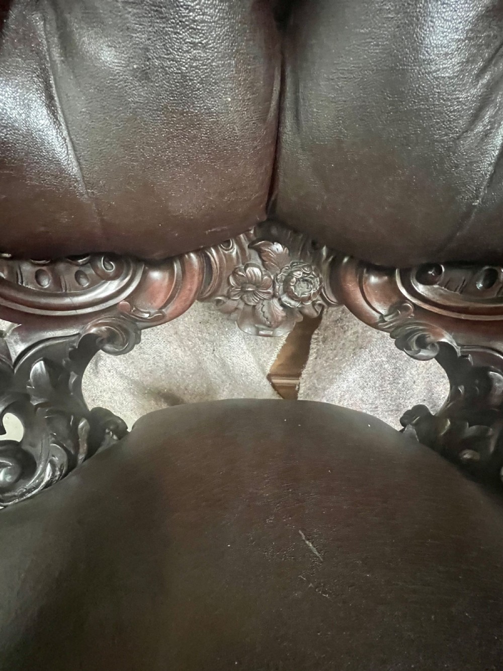 A Victorian carved mahogany armchair featuring deep-buttoned brown leather upholstery, elaborate scrollwork carving, carved armrests, and cabriole legs with brass castors. View XIV