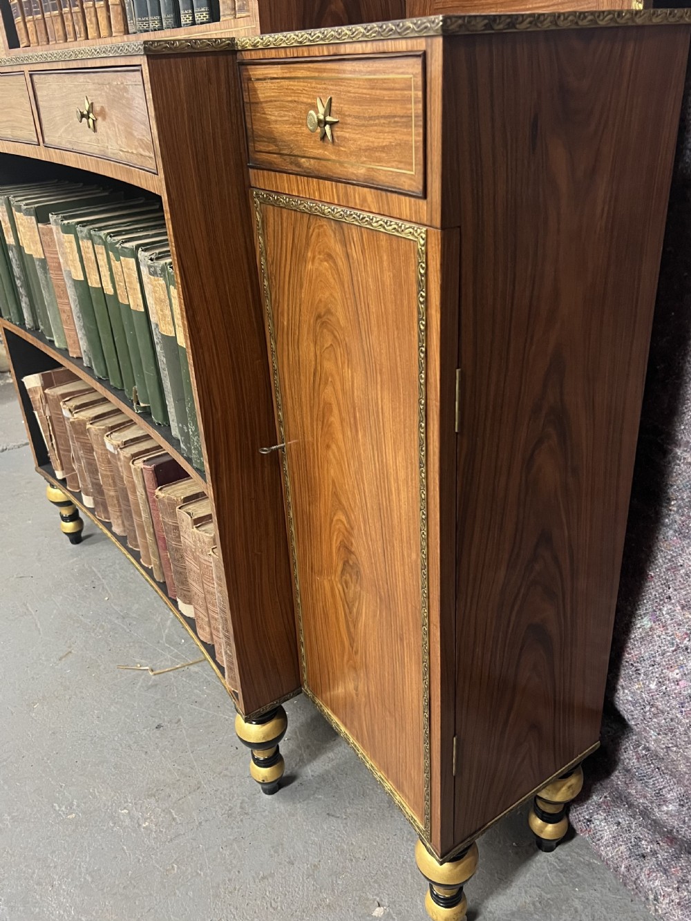 A Regency style rosewood breakfront open bookcase with tiered shelving filled with books, brass star handles, mirrored side compartments, and turned legs. View VIII