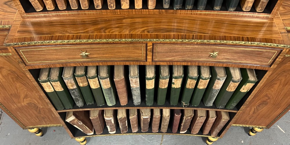 A Regency style rosewood breakfront open bookcase with tiered shelving filled with books, brass star handles, mirrored side compartments, and turned legs. View X