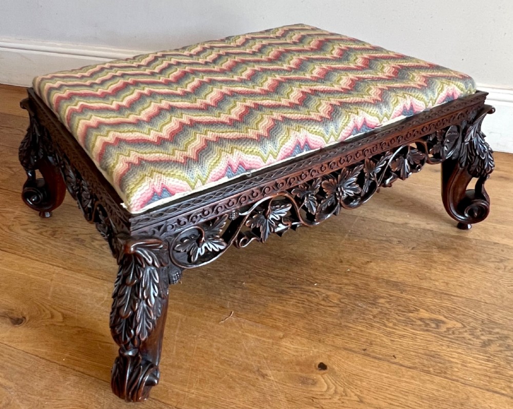 A large rectangular Anglo-Indian stool featuring an intricately carved dark hardwood apron and cabriole legs, topped with a flame-stitch patterned tapestry cushion.