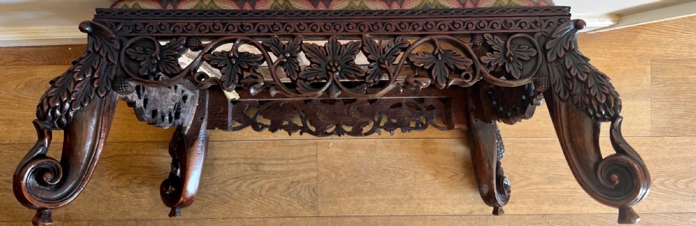 A large rectangular Anglo-Indian stool featuring an intricately carved dark hardwood apron and cabriole legs, topped with a flame-stitch patterned tapestry cushion. View VI