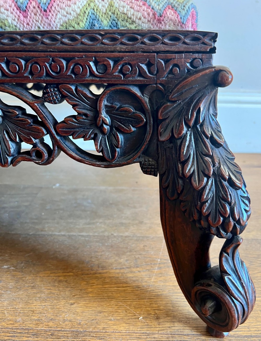 A large rectangular Anglo-Indian stool featuring an intricately carved dark hardwood apron and cabriole legs, topped with a flame-stitch patterned tapestry cushion. View VII