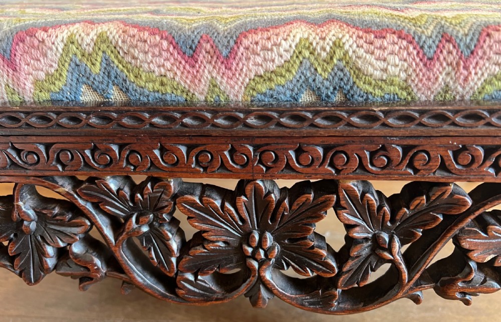 A large rectangular Anglo-Indian stool featuring an intricately carved dark hardwood apron and cabriole legs, topped with a flame-stitch patterned tapestry cushion. View VIII