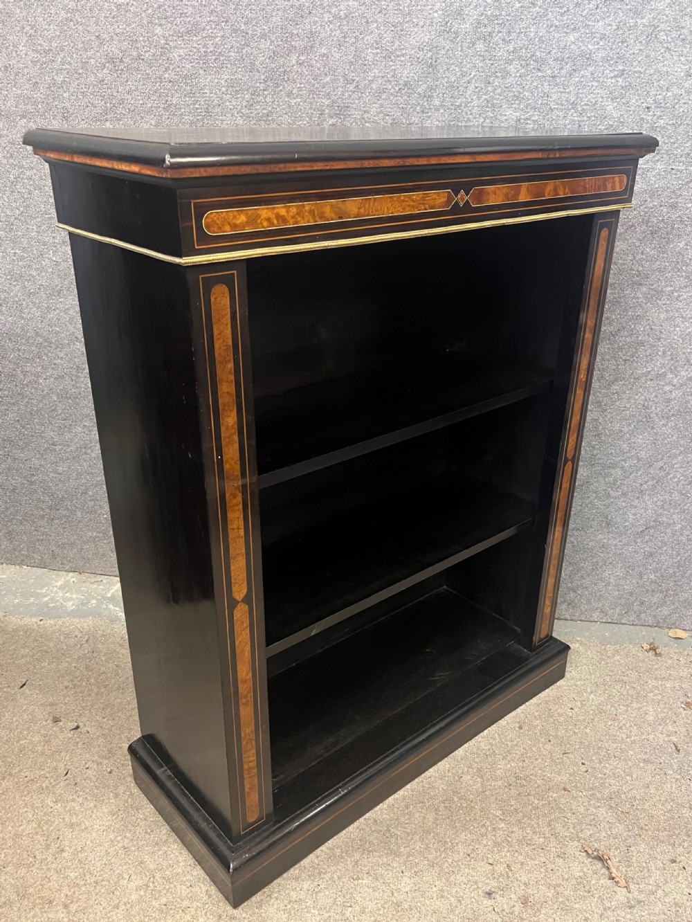 A small ebonised open bookcase with three shelves, featuring burr walnut inlay banding and thin brass moulding detailing.