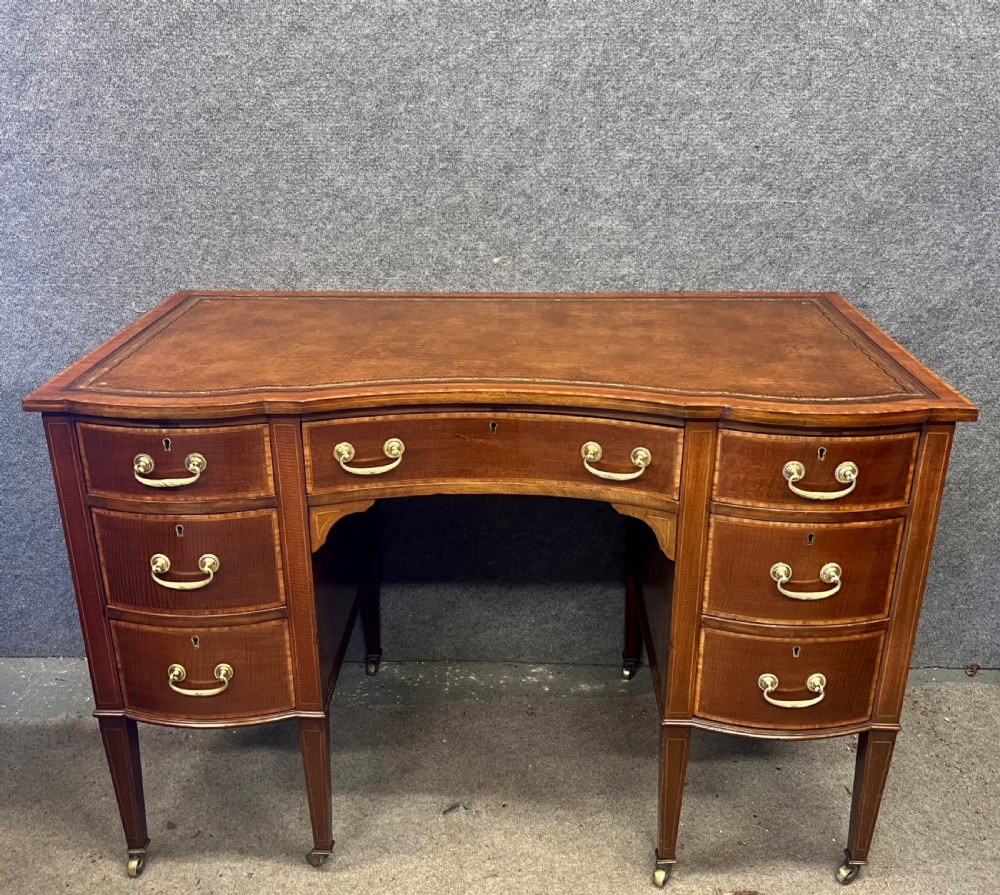An Edwardian inlaid mahogany writing desk featuring a leather-lined top, a curved serpentine front, seven drawers with brass drop handles, and tapered square legs on castors.