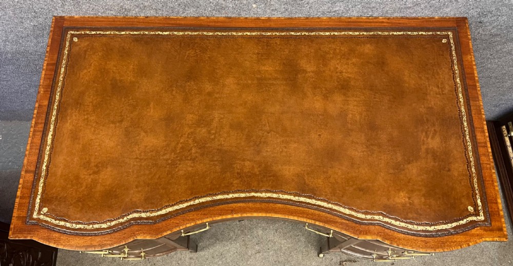 An Edwardian inlaid mahogany writing desk featuring a leather-lined top, a curved serpentine front, seven drawers with brass drop handles, and tapered square legs on castors. View II
