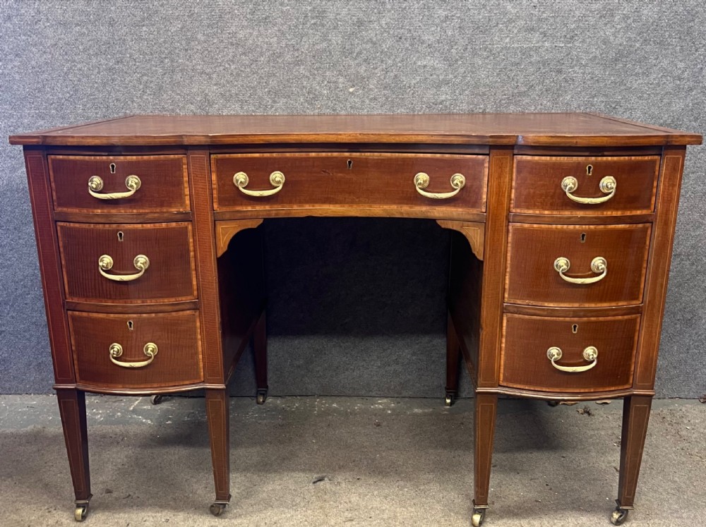 An Edwardian inlaid mahogany writing desk featuring a leather-lined top, a curved serpentine front, seven drawers with brass drop handles, and tapered square legs on castors. View III