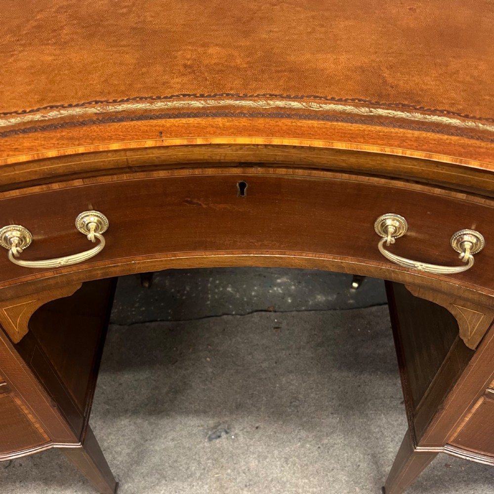 An Edwardian inlaid mahogany writing desk featuring a leather-lined top, a curved serpentine front, seven drawers with brass drop handles, and tapered square legs on castors. View VII
