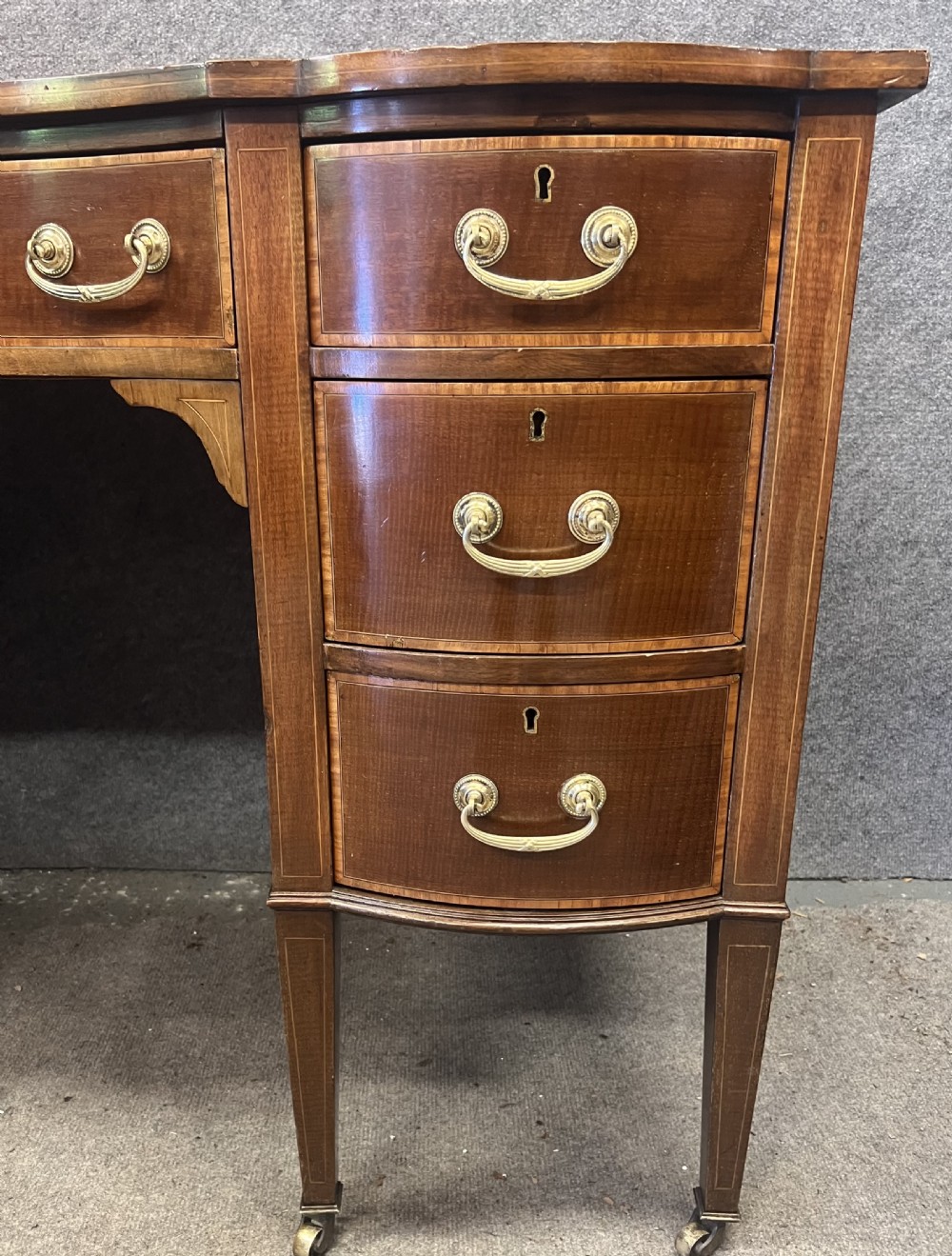 An Edwardian inlaid mahogany writing desk featuring a leather-lined top, a curved serpentine front, seven drawers with brass drop handles, and tapered square legs on castors. View VIII