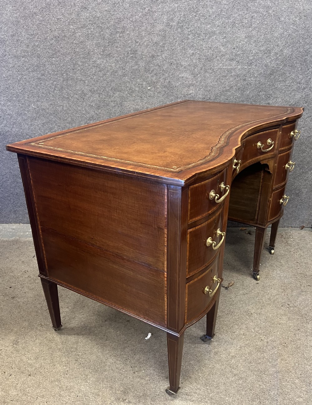 An Edwardian inlaid mahogany writing desk featuring a leather-lined top, a curved serpentine front, seven drawers with brass drop handles, and tapered square legs on castors. View X