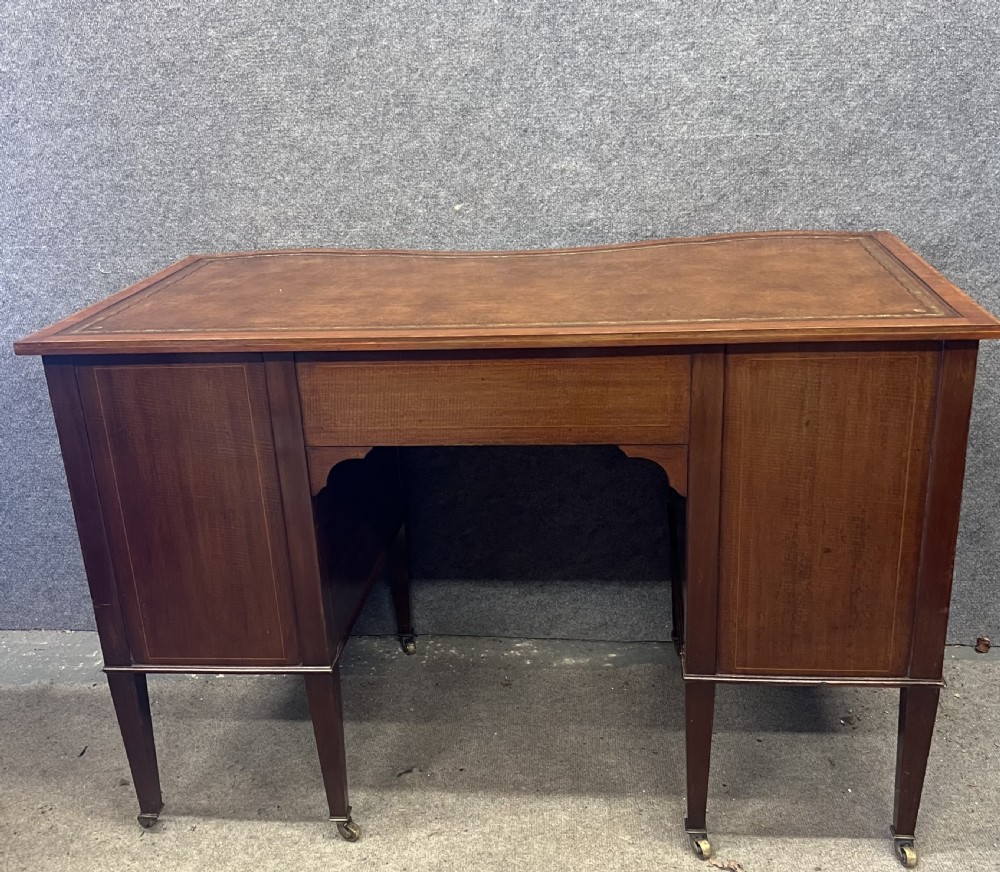 An Edwardian inlaid mahogany writing desk featuring a leather-lined top, a curved serpentine front, seven drawers with brass drop handles, and tapered square legs on castors. View XI