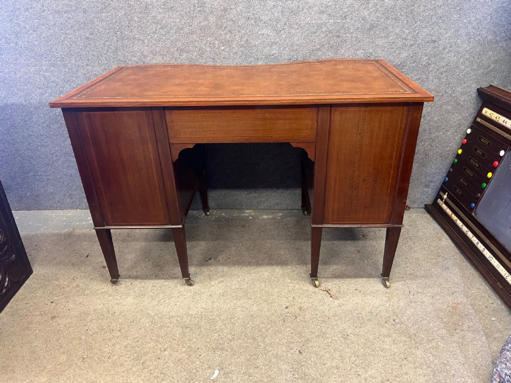 An Edwardian inlaid mahogany writing desk featuring a leather-lined top, a curved serpentine front, seven drawers with brass drop handles, and tapered square legs on castors. View XIV
