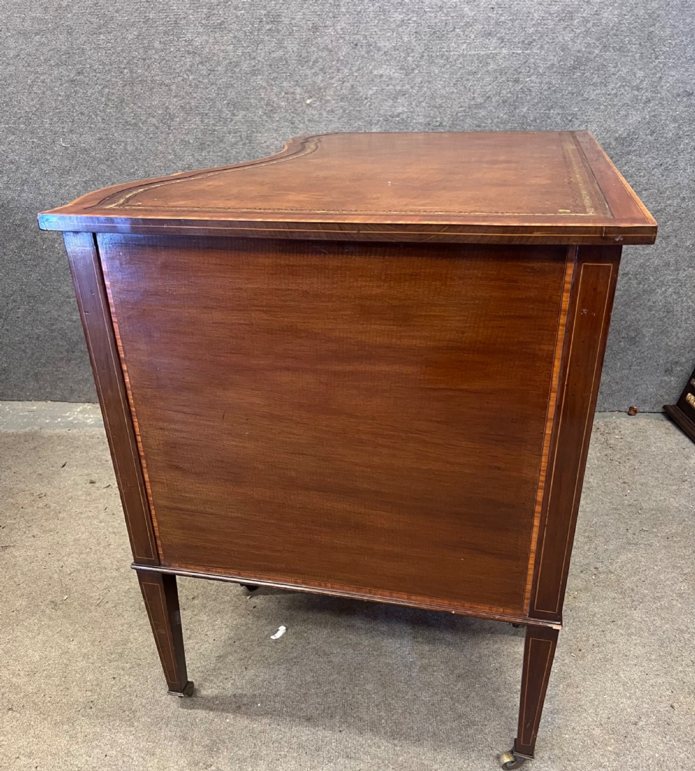 An Edwardian inlaid mahogany writing desk featuring a leather-lined top, a curved serpentine front, seven drawers with brass drop handles, and tapered square legs on castors. View XVI