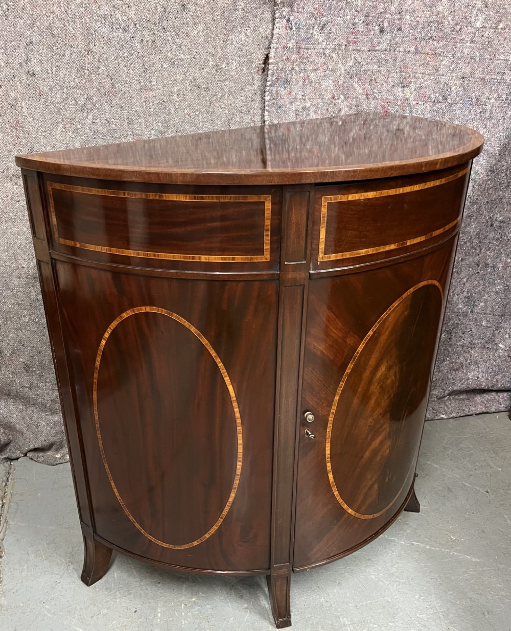 A semi-circular Sheraton Revival mahogany cabinet featuring decorative oval wood inlays, stringing details, and square tapered legs.