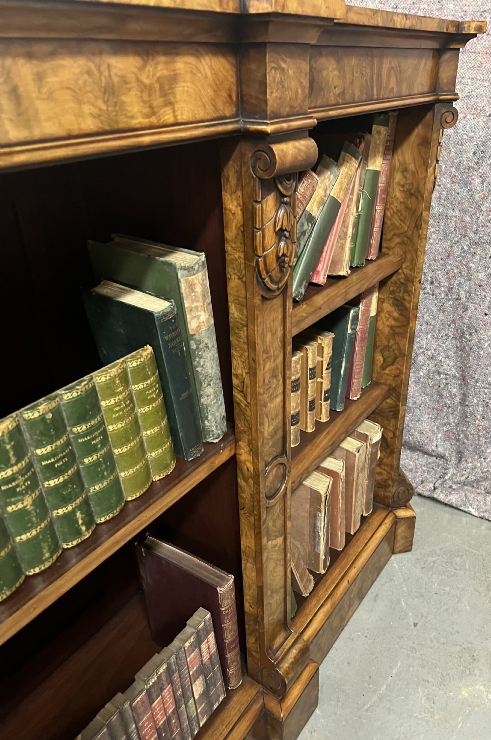 A low, wide Victorian burr walnut open bookcase with three compartmentalised shelf sections, carved pilaster corbels, and a solid plinth base. View XI
