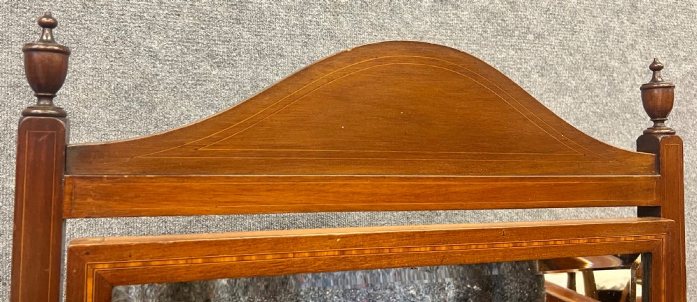 An Edwardian mahogany cheval mirror featuring an arched cresting rail with satinwood line inlay, turned brass finials, bevelled glass plate, and splayed legs on castors. View V