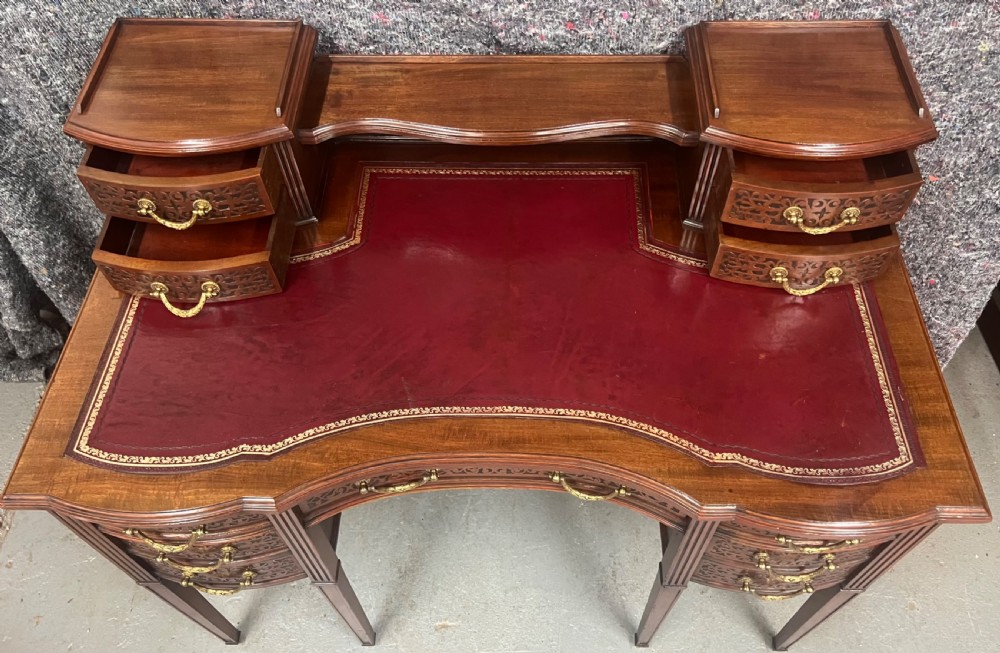 A mahogany writing desk featuring a red leather inset surface, carved drawer fronts, brass swanneck handles, and an elevated superstructure with twin drawer units. View V